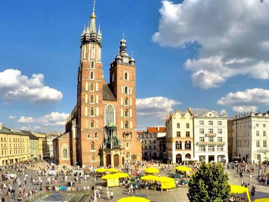 St. Mary's Basilica in Krak&oacute;w's main square, with crowds of people, yellow market stalls, and historic buildings under a partly cloudy sky.