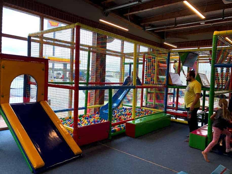 Indoor playground area with a slide, climbing structures, and a ball pit at Majaland Amusement Park Kownaty. An adult stands nearby while children play inside the enclosed play equipment.