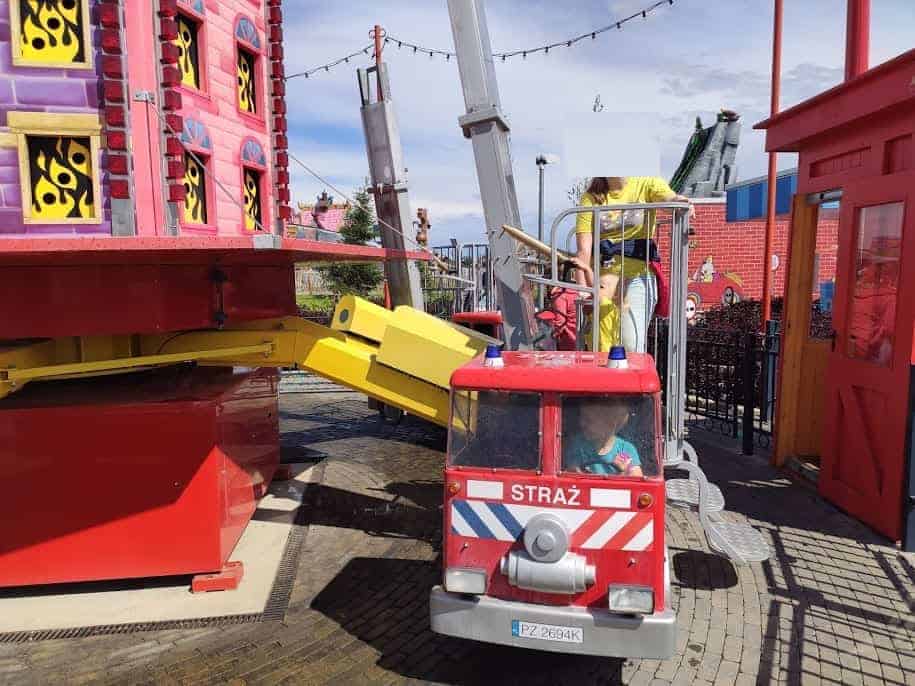 A child enjoys a small fire truck ride labeled "STRAŻ" at Majaland Kownaty, an amusement park with colorful buildings and a raised mechanical platform.