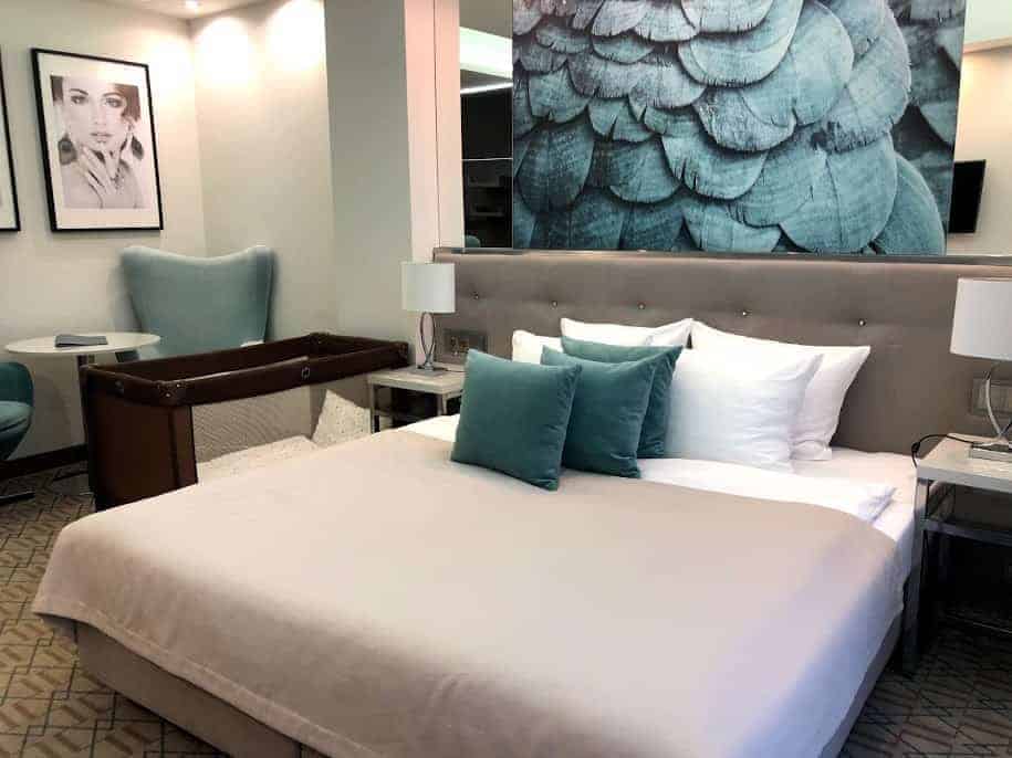 Modern hotel room with a double bed, teal accent pillows, two bedside tables, a crib, armchair, and framed wall art&mdash;perfect for guests who want to relax after exploring Poland using helpful travel apps.
