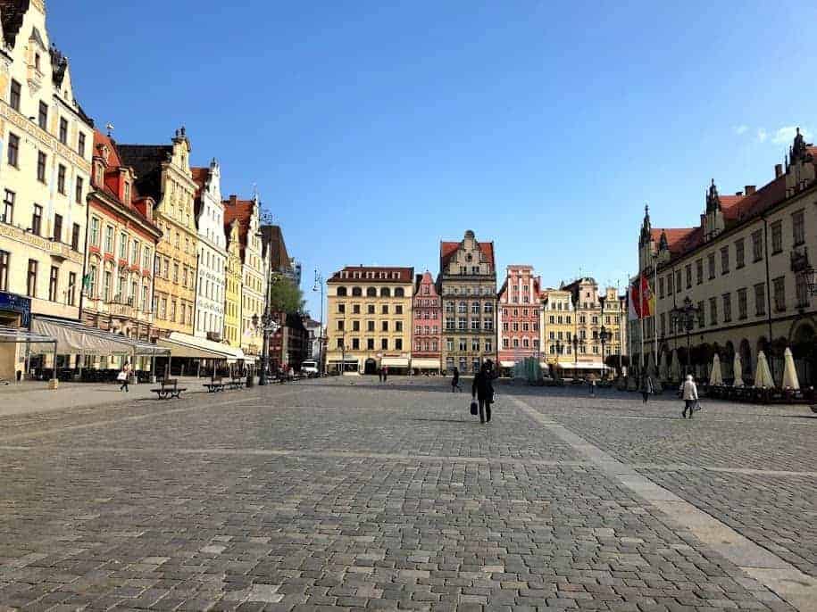 A spacious cobblestone square in Dolnośląskie, Poland, lined with colorful historic buildings under a clear blue sky; a few people walk in the open area&mdash;one of the best places to visit.