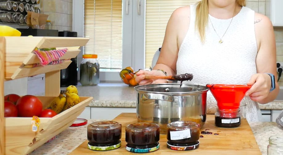 A person in a kitchen is using a ladle to pour a dark liquid from a pot through a red funnel into a jar, likely following an old Polish Strawberry Jam recipe, with other jars and fresh produce visible on the countertop.