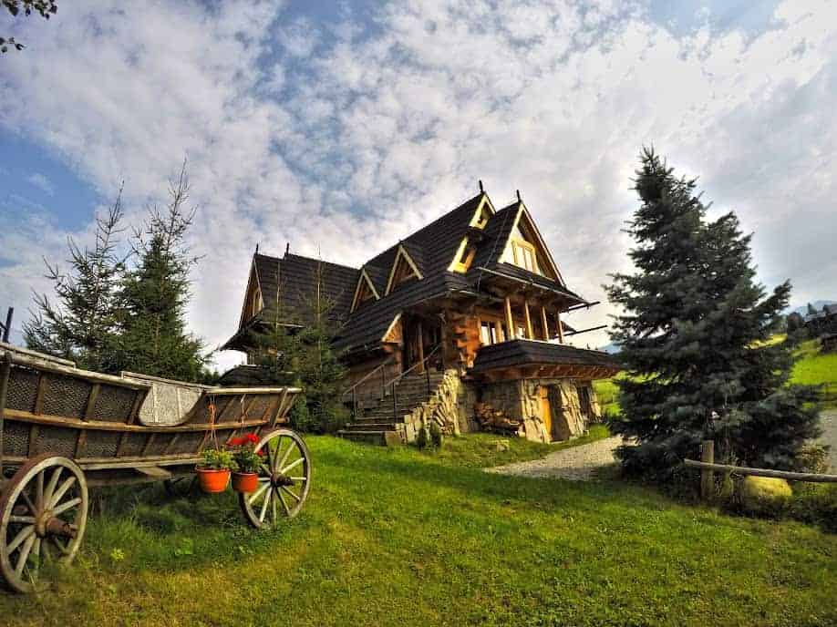 A rustic wooden house with a steep roof sits on a grassy lawn, next to an old wooden cart and pine trees, under a partly cloudy sky.