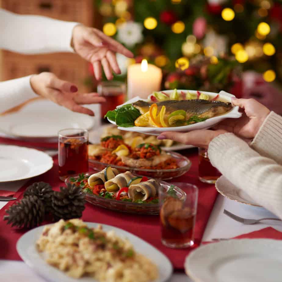 A holiday table comes alive with Polish flavors as people pass a platter of fish, surrounded by diverse dishes, drinks, and a lit candle. A beautifully decorated Christmas tree twinkles in the background, making it a perfect gathering for foodies.