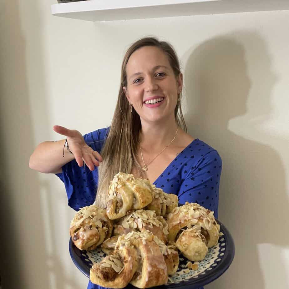 A woman in a blue shirt holds a plate piled with freshly baked Rogale Świętomarcińskie, topped with almonds, smiling at the camera.