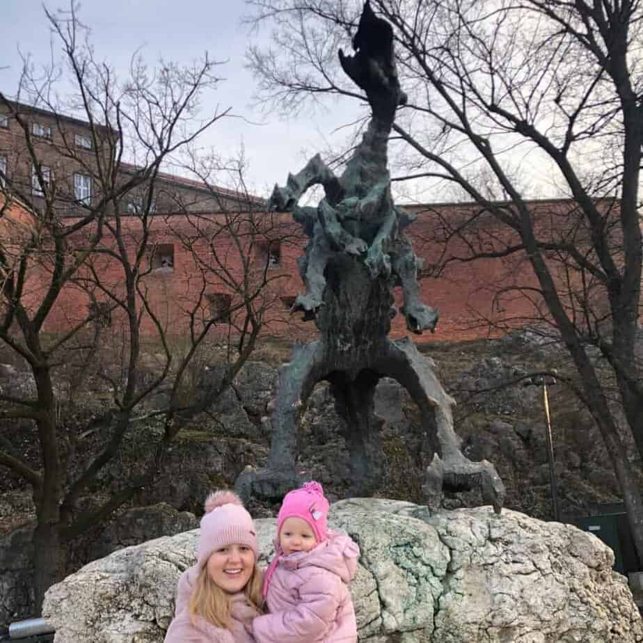 A woman and a child in pink winter clothing stand before a large dragon statue on a rock&mdash;one of the top Krak&oacute;w attractions&mdash;with bare trees and a brick building in the background.
