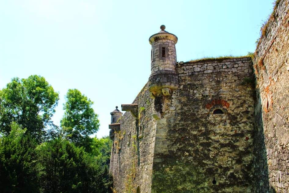 Stone fortress wall with cylindrical watchtowers, surrounded by trees and greenery under a clear blue sky&mdash;a stunning example of must-see castles in Poland.