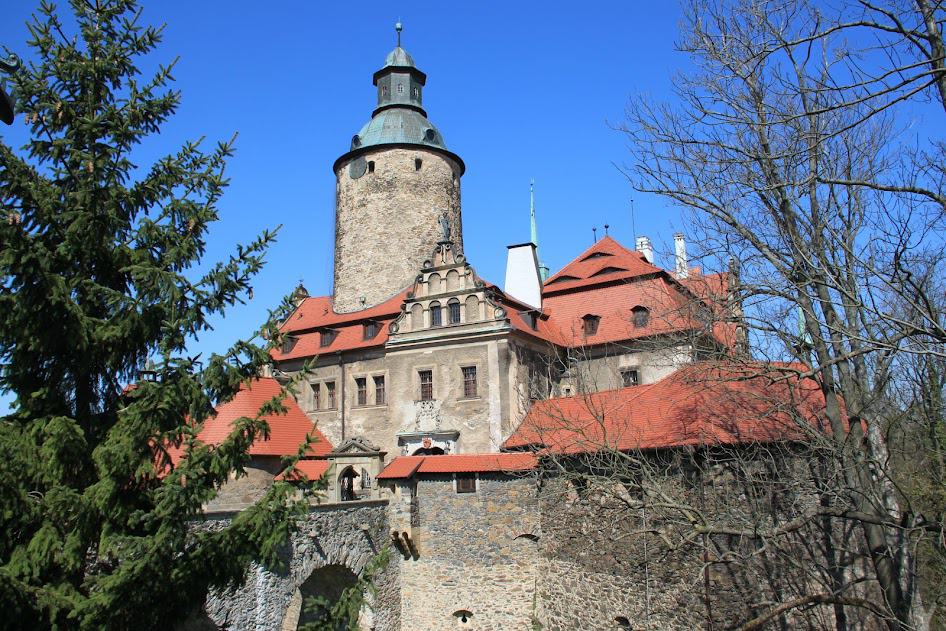 A large stone castle with a round tower, red-tiled roofs, and arched windows, surrounded by trees under a clear blue sky&mdash;one of the must-see castles in Poland for any Poland tourism enthusiast.