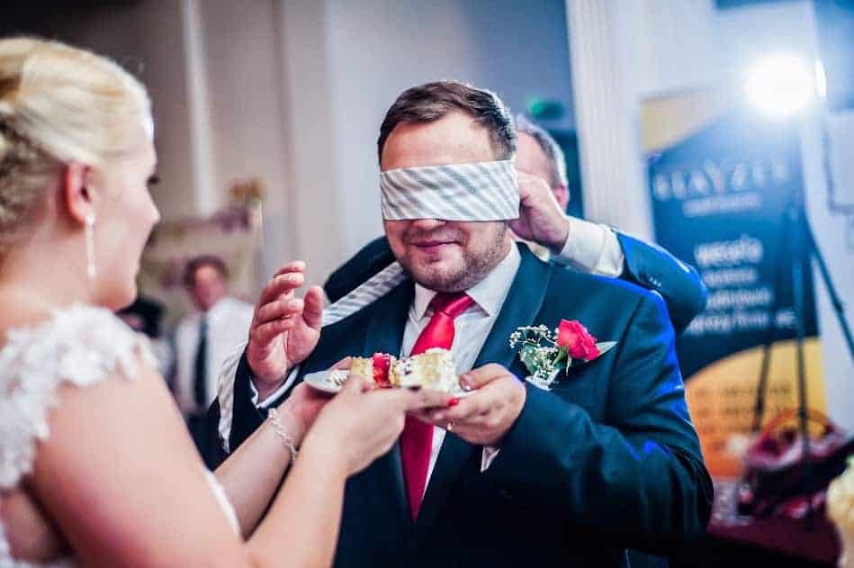 A man in a suit with a blindfold is being offered a piece of cake by a woman in a wedding dress at a celebration, reflecting playful Polish wedding traditions.