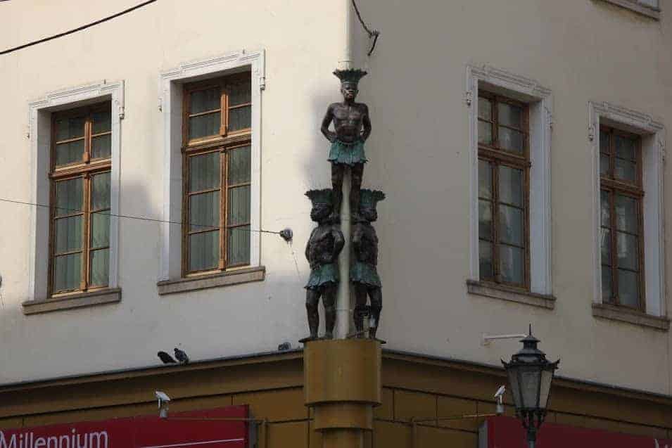 A sculpture of three standing male figures, stacked vertically, is mounted on the corner of a building with white walls and wooden-framed windows&mdash;a striking example of Wroclaw Street Art.