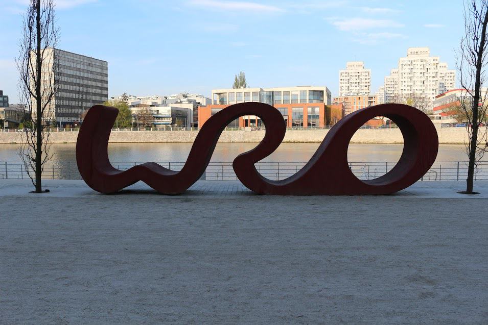 Large red metal sculpture resembling the letters "WRO" stands in an outdoor urban area near a river, showcasing Wroclaw Street Art with modern buildings in the background.
