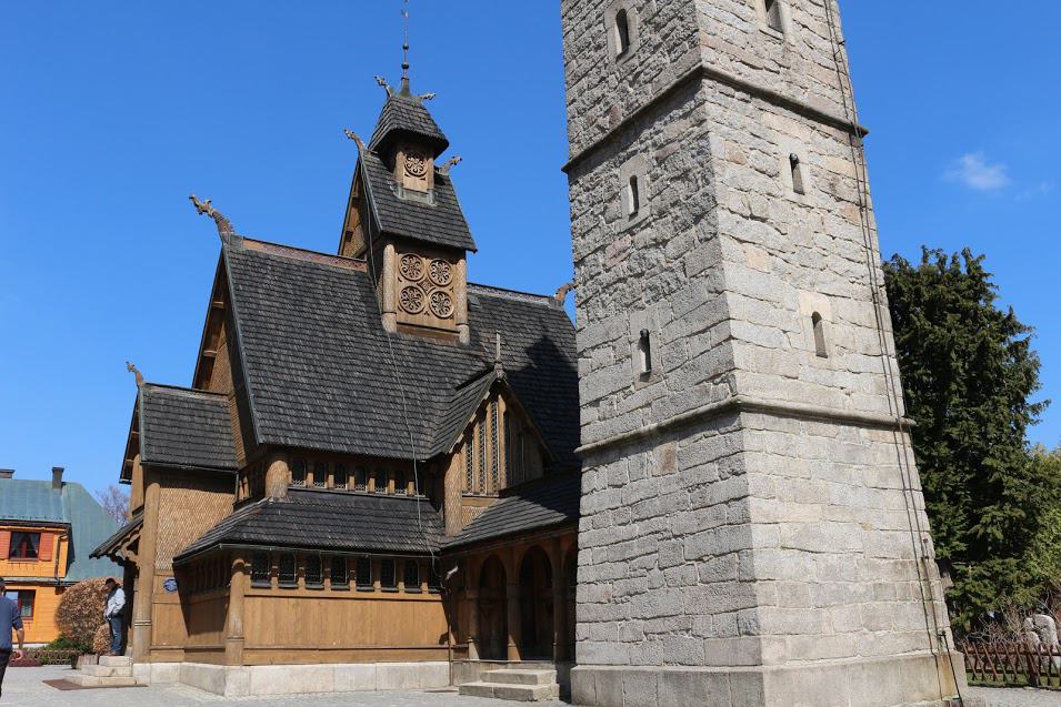 A wooden stave church with ornate carvings stands beside a tall stone bell tower under a clear blue sky&mdash;one of the best places in Dolnośląskie Poland to explore history and architecture.