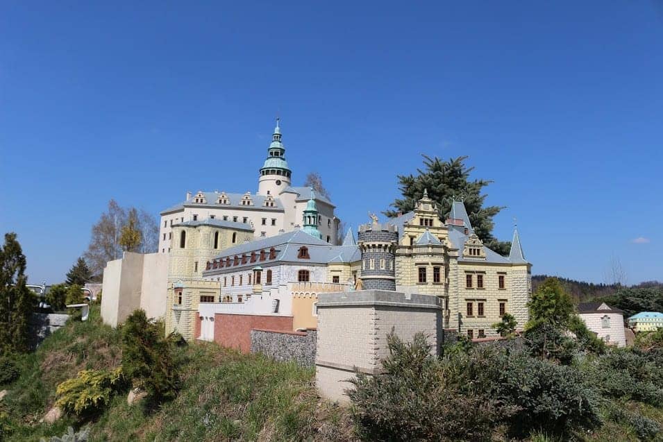 A detailed model of a European-style castle with multiple towers and spires, inspired by some of the best places to visit in Dolnośląskie, Poland, and surrounded by shrubbery under a clear blue sky.