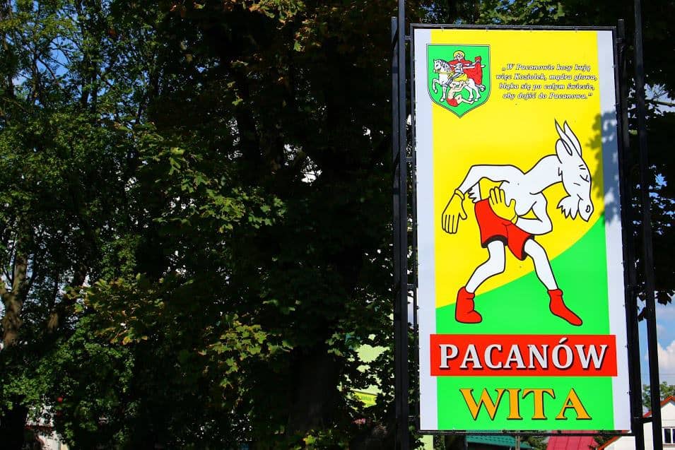 Sign with a cartoon goat wearing red shorts and a yellow shirt, the words "Pacanów Wita," and a coat of arms on a green and yellow background. A fun highlight in Swietokrzyskie, Poland—one of the charming places to visit. Trees in the background.