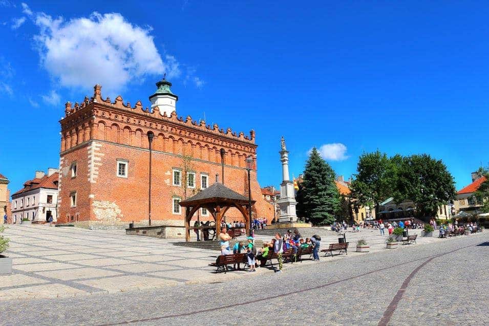 A historic brick building with a clock tower stands in a sunny town square in Swietokrzyskie, Poland—one of the best places to visit—surrounded by people on benches and strolling past trees and smaller buildings in the background.