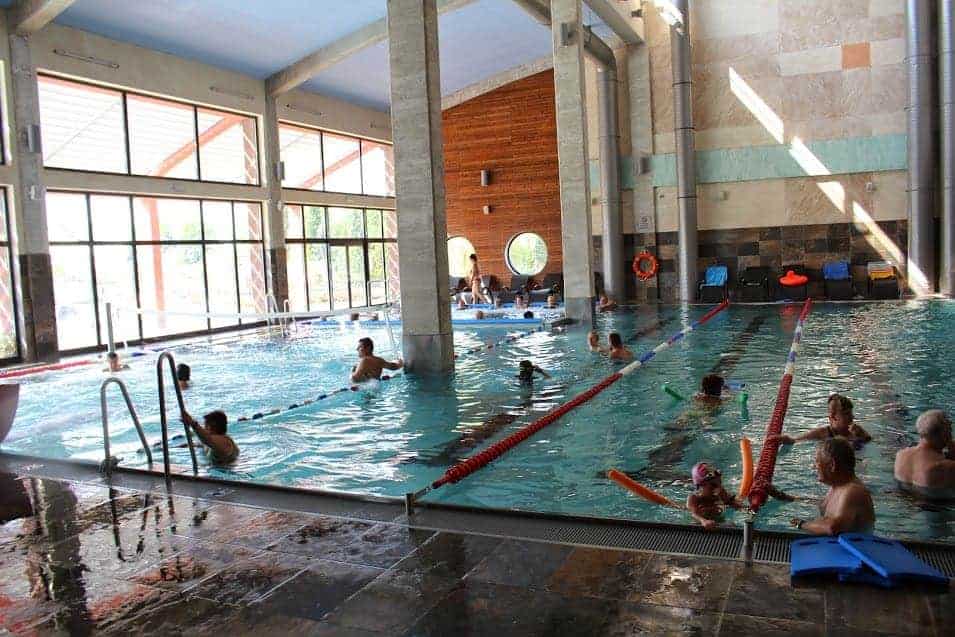 Indoor swimming pool in Swietokrzyskie, Poland, with several people, including children and adults, swimming and playing. Sunlight enters through large windows on the left. Pool equipment is visible on the deck—one of the great places to visit.