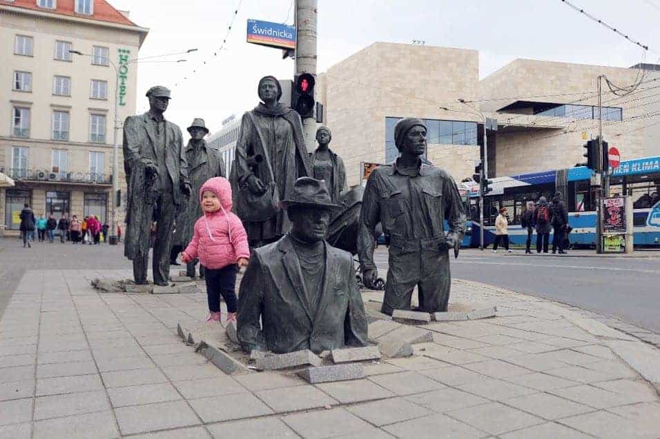 A young child in a pink jacket stands among life-size bronze statues of people partially sinking into the sidewalk on a city street corner, capturing a striking scene of Street Art Wroclaw.