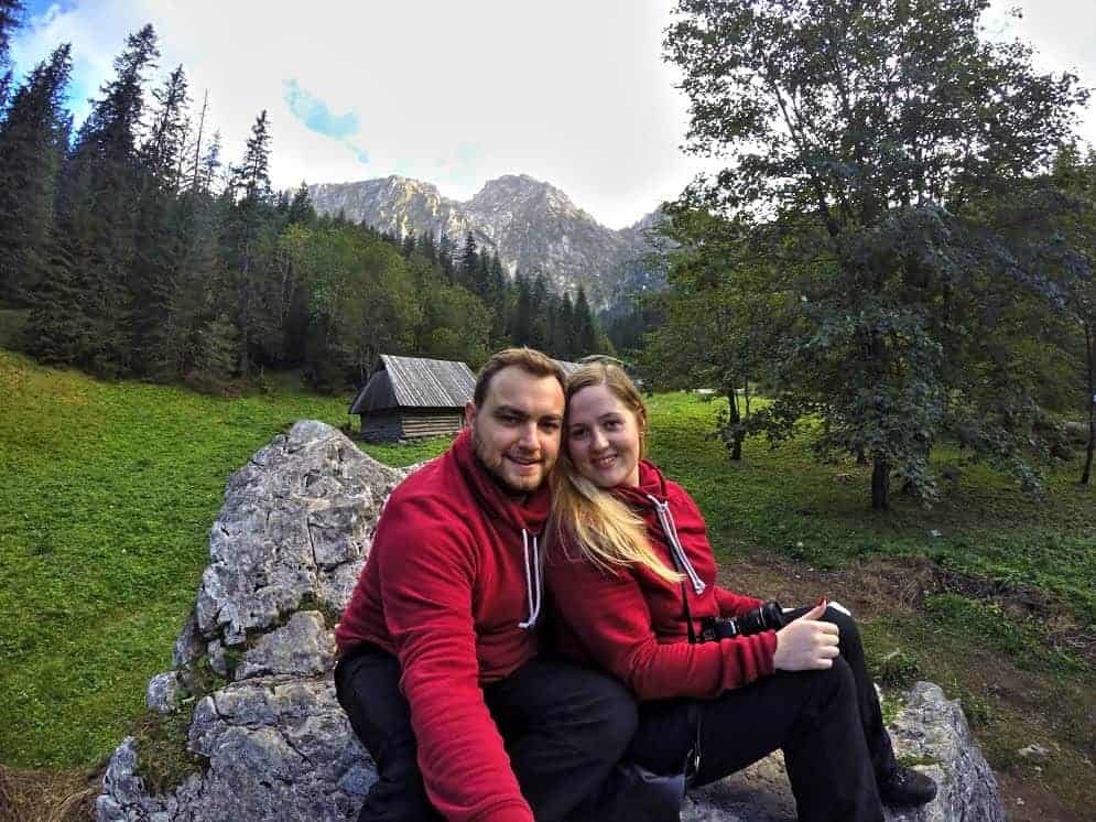 A man and a woman in matching red hoodies sit on a rock in a green, mountainous landscape with trees and a small cabin&mdash;one of the many romantic places in Poland perfect for couples to escape together.