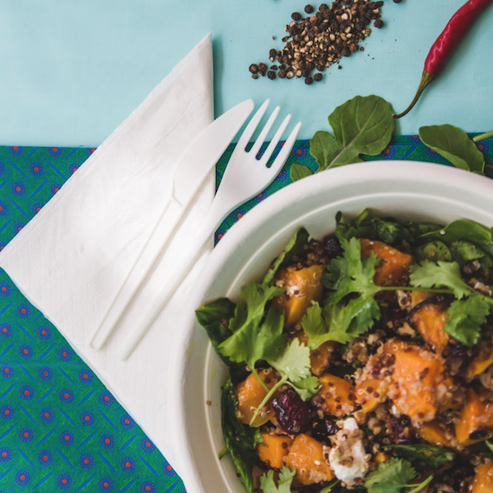 A folded serviette with a CPLA knife and fork on top, next to a sugarcane bowl filled with roast butternut salad
