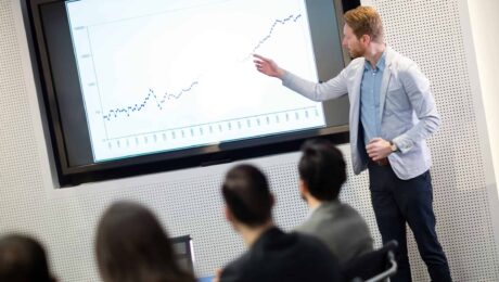 a man standing in front of a screen pointing at a graph