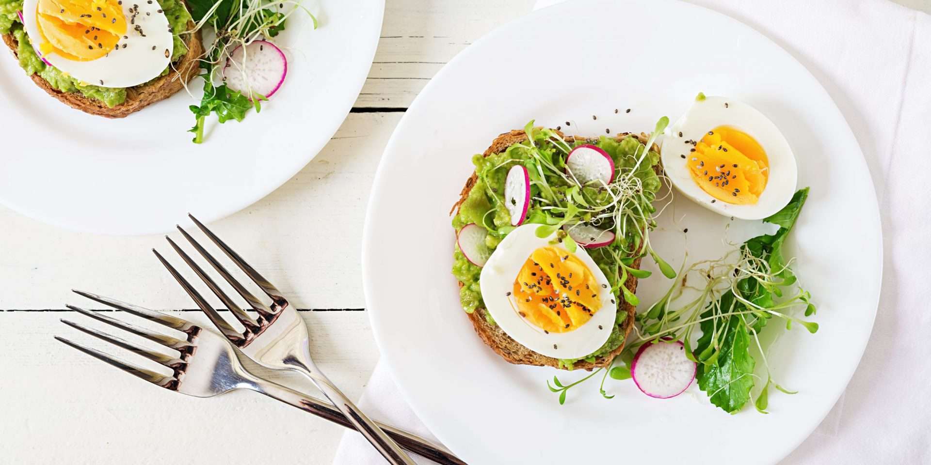 Open-faced sandwiches topped with avocado, radishes, greens, and a boiled egg on white plates, inspired by naturopathic medicine principles.