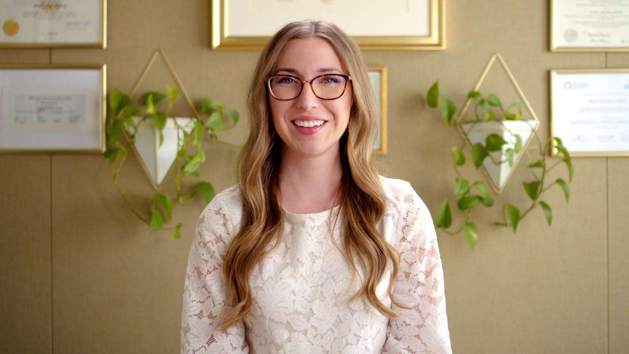 Dr. Alexa NMD standing in front of a wall adorned with framed certificates and decorative plants, showcasing her expertise in naturopathic medicine.