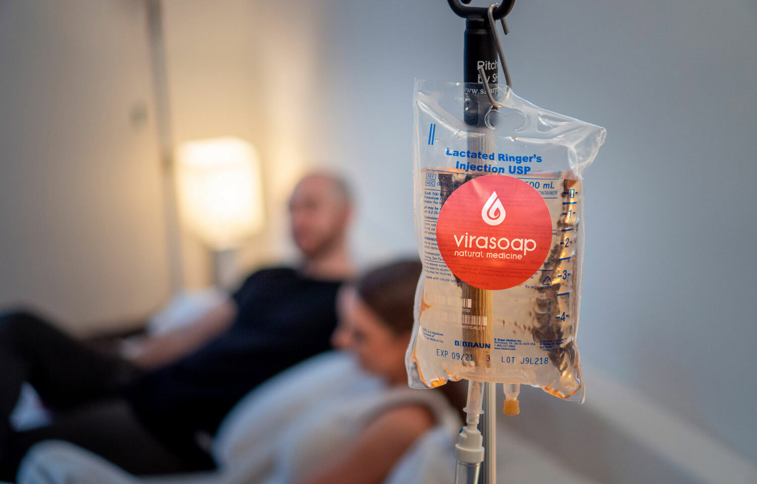 A close-up of an IV bag labeled "lactated ringer's injection USP" with the Virasoap Natural Medicine logo in a medical setting, with a blurred background showing a reclining man and woman suffering from fatigue in Meridian, Idaho
