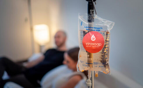 A close-up of an IV bag labeled "lactated ringer's injection USP" with the Virasoap Natural Medicine logo in a medical setting, with a blurred background showing a reclining man and woman suffering from fatigue in Meridian, Idaho