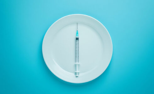 A syringe, representing GLP-1 meds, is placed on a white plate against a blue background.