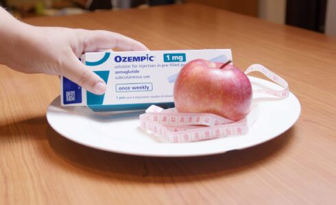 A hand places an Ozempic 1mg box, a natural GLP-1 medication, next to a red apple and measuring tape on a white plate on a wooden table.