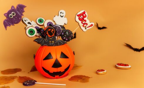 A Halloween-themed arrangement with decorated lollipops in a pumpkin container. Lollipops include eyes, ghosts, bats, and a "Boo!" sign. Background features bats and fallen leaves.