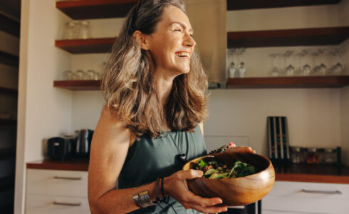 Aging woman smiling happily while holding a buddha bowl in her kitchen. Happy middle aged woman serving herself a healthy vegan meal at home, taking care of her body with a plant-based diet.