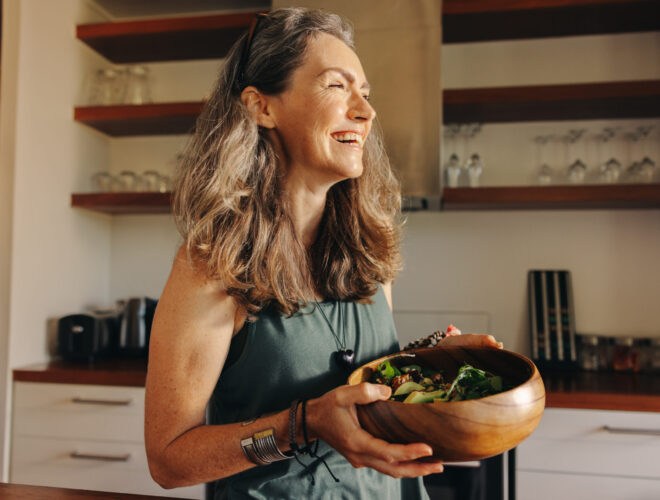 Aging woman smiling happily while holding a buddha bowl in her kitchen. Happy middle aged woman serving herself a healthy vegan meal at home, taking care of her body with a plant-based diet.