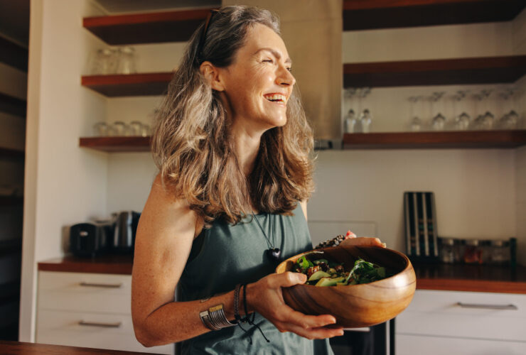 Aging woman smiling happily while holding a buddha bowl in her kitchen. Happy middle aged woman serving herself a healthy vegan meal at home, taking care of her body with a plant-based diet.