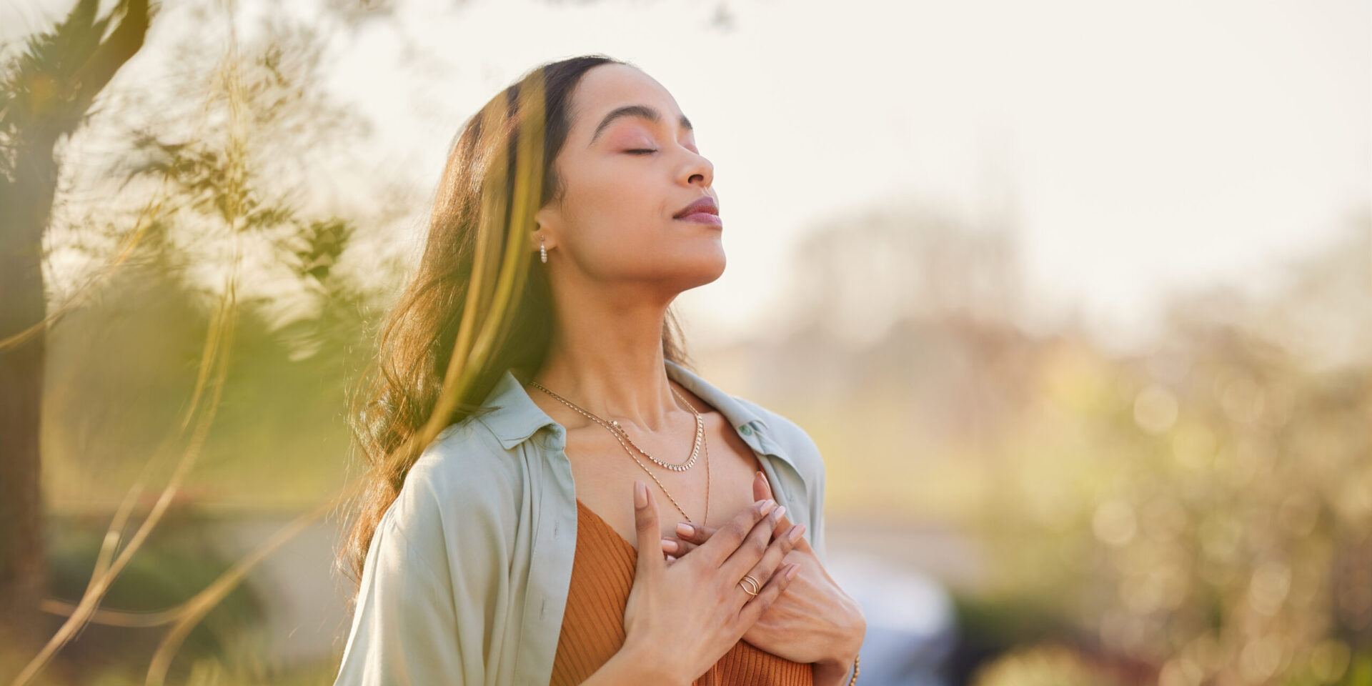 A woman stands outside with her eyes closed and hands on her chest, appearing calm and relaxed in a sunlit, natural setting—embracing tranquility as part of her cholesterol management journey.