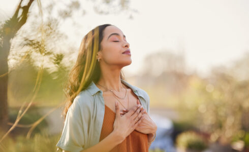 A woman stands outside with her eyes closed and hands on her chest, appearing calm and relaxed in a sunlit, natural setting—embracing tranquility as part of her cholesterol management journey.