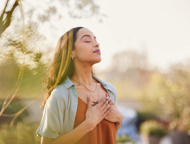 A woman stands outside with her eyes closed and hands on her chest, appearing calm and relaxed in a sunlit, natural setting—embracing tranquility as part of her cholesterol management journey.