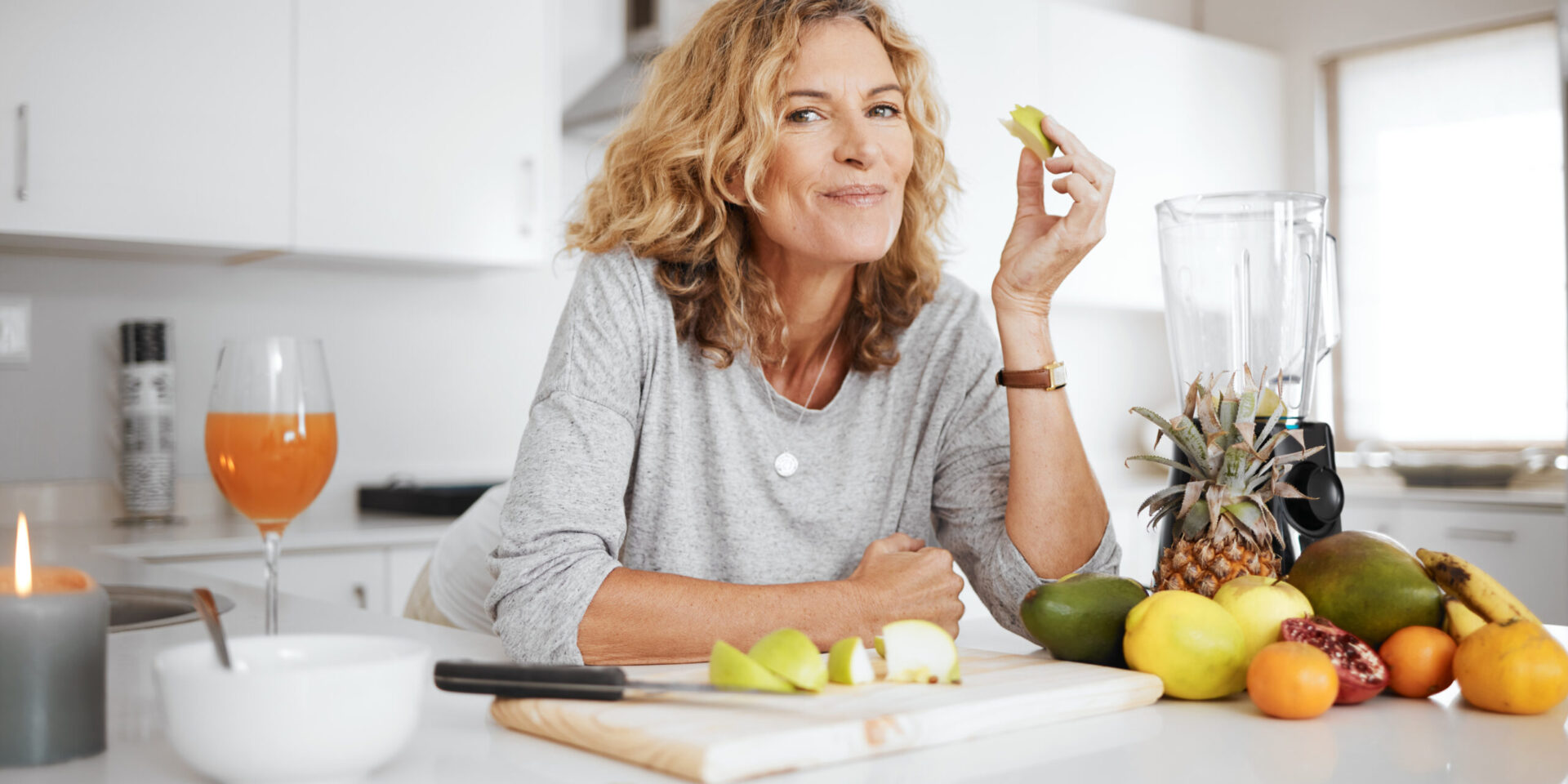 Woman in a kitchen smiling while holding a slice of green apple, surrounded by fruits, a blender, and a glass of juice—creating the perfect setting to improve your diet and nourish your skin.