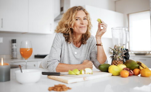 Woman in a kitchen smiling while holding a slice of green apple, surrounded by fruits, a blender, and a glass of juice—creating the perfect setting to improve your diet and nourish your skin.