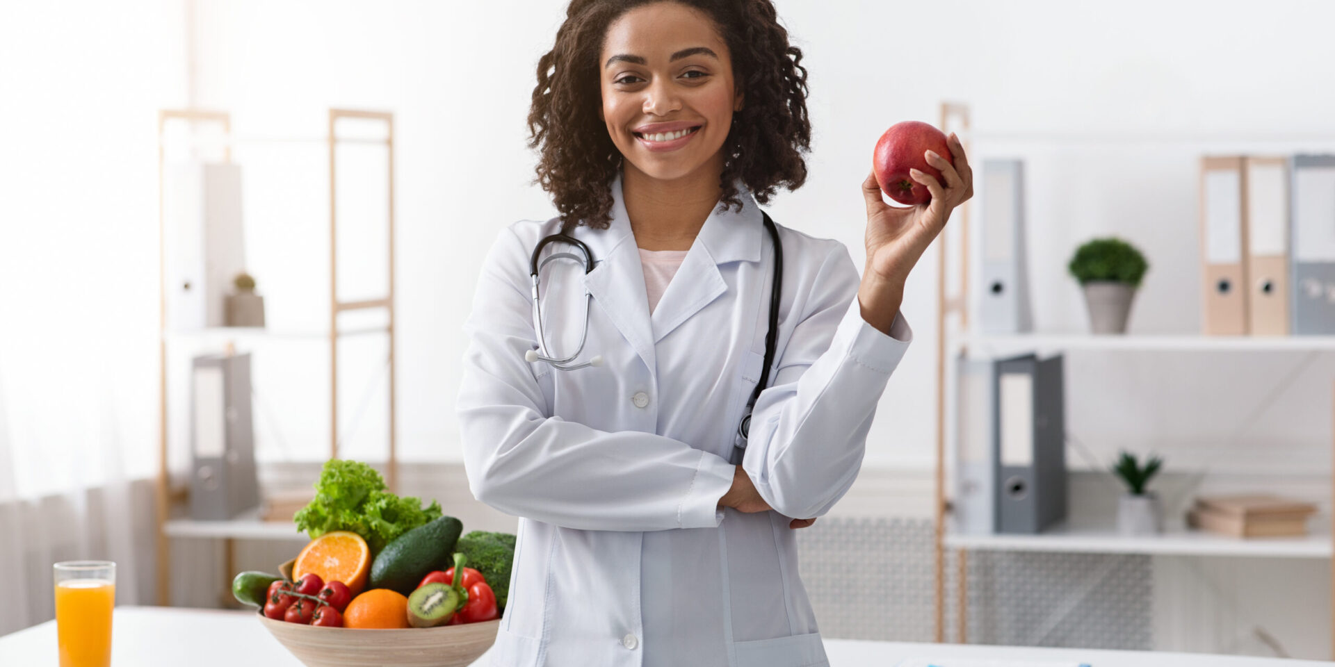 A doctor wearing a white coat and stethoscope stands smiling, holding an apple; a bowl of assorted fruits and vegetables is on the table beside her, highlighting the hormone connection through nutrition and gut health.