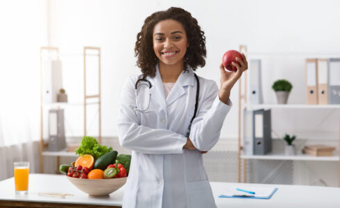 A doctor wearing a white coat and stethoscope stands smiling, holding an apple; a bowl of assorted fruits and vegetables is on the table beside her, highlighting the hormone connection through nutrition and gut health.