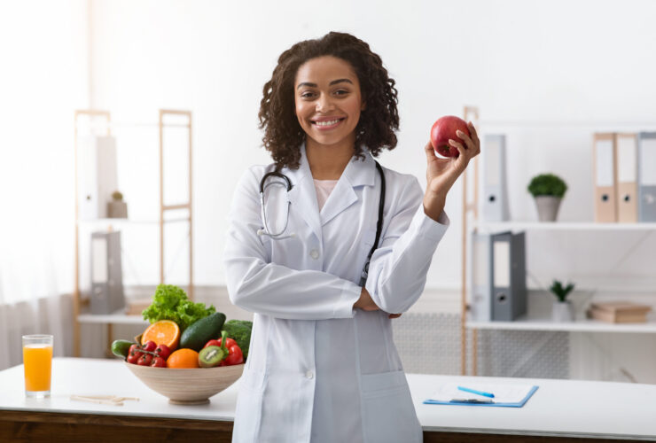 A doctor wearing a white coat and stethoscope stands smiling, holding an apple; a bowl of assorted fruits and vegetables is on the table beside her, highlighting the hormone connection through nutrition and gut health.