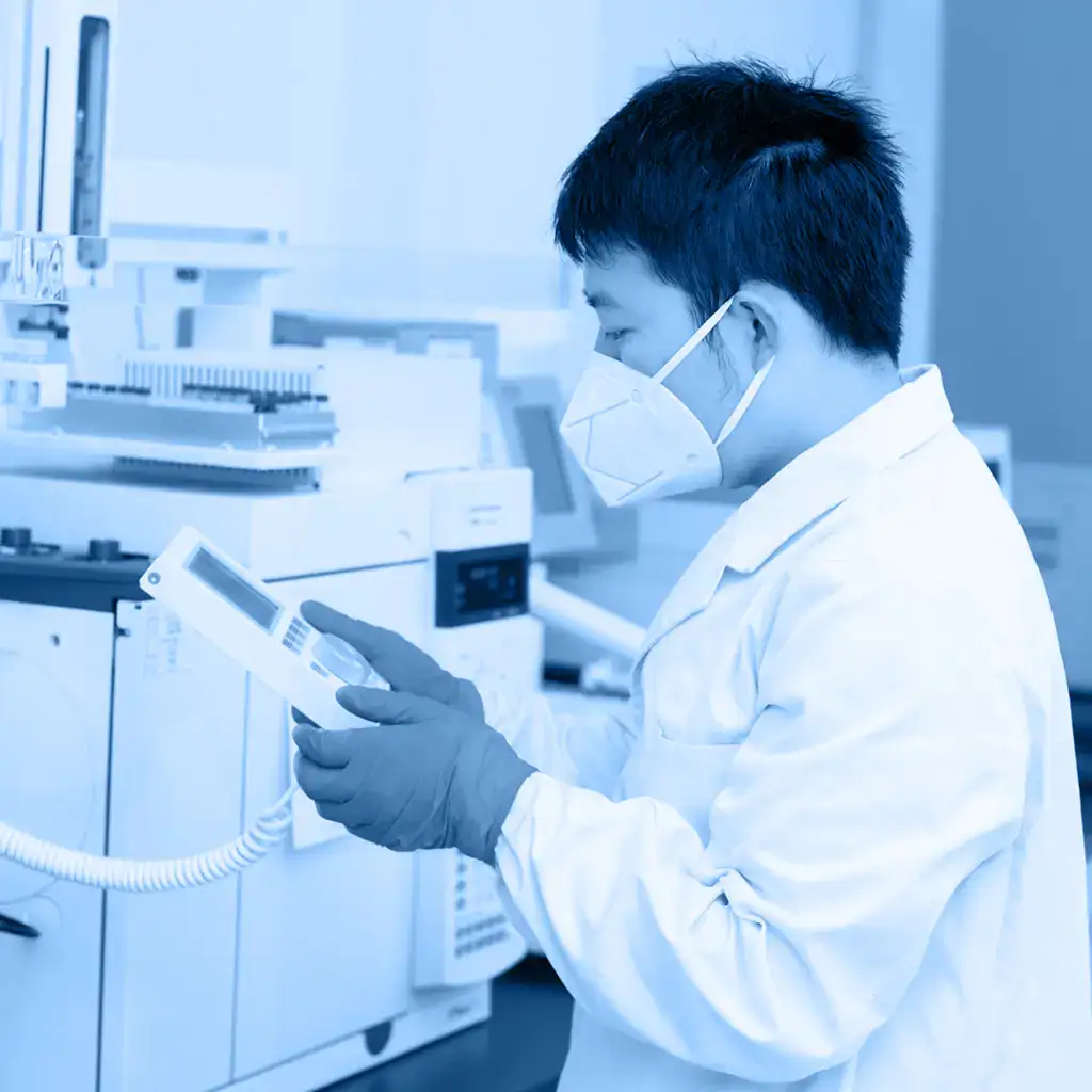 Laboratory technician in a white lab coat and mask reviewing results on a handheld device with medical equipment in the background.