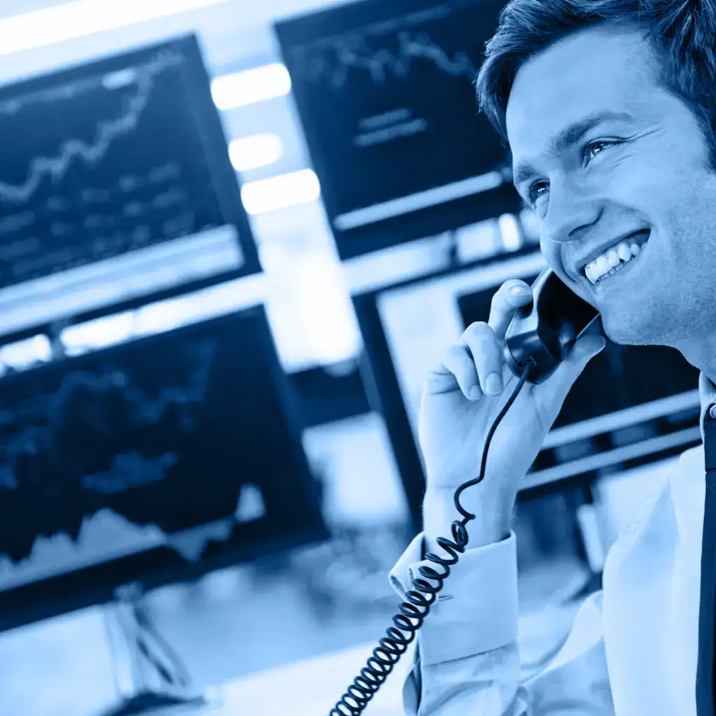 Cheerful financial analyst talking on the phone with stock market data on multiple monitors in the background.
