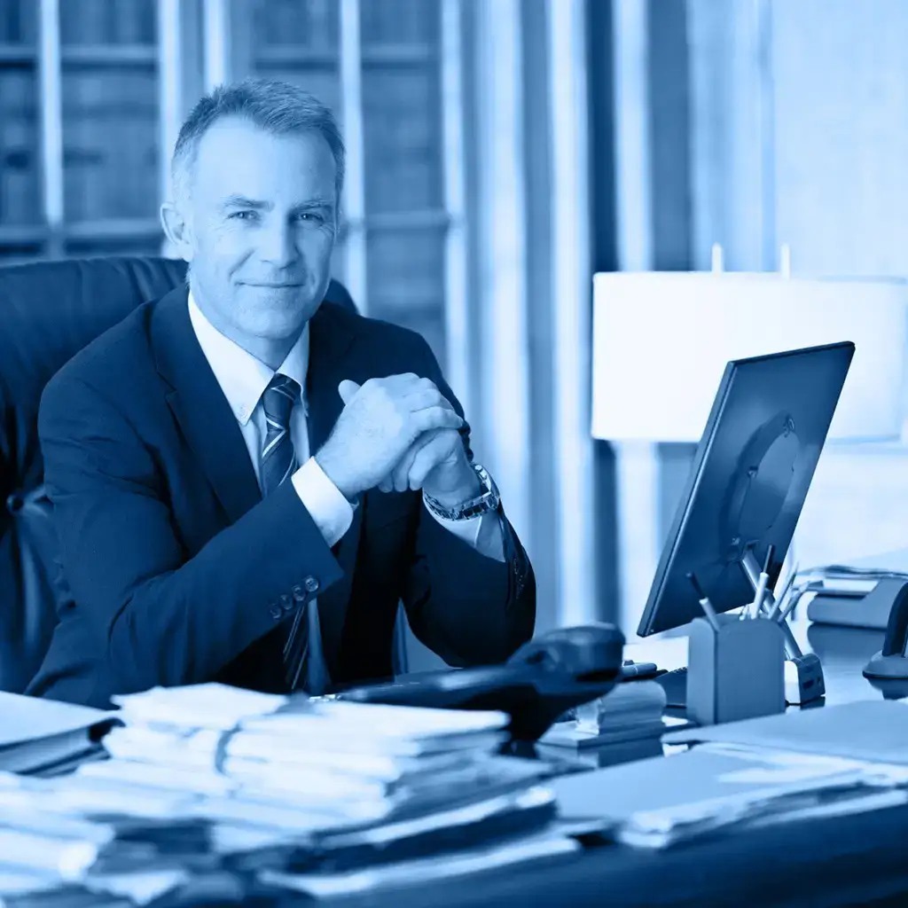 Senior businessman in a blue-tinted office, looking thoughtful, with hands clasped and a laptop on the desk.