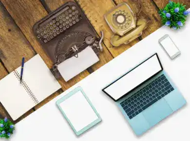 shutterstock_1084997315 - ATECHSO - Managed IT Company Top-down view of a vintage typewriter, rotary phone, and modern laptop and smartphone on a wooden desk, contrasting old and new communication technologies.