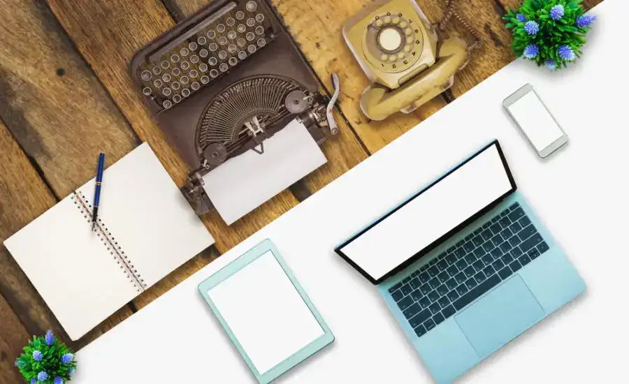 Top-down view of a vintage typewriter, rotary phone, and modern laptop and smartphone on a wooden desk, contrasting old and new communication technologies.